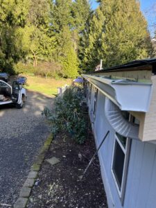 A close-up view of a house gutter system with downspout, next to a gravel driveway, shrubs, and a parked vehicle, surrounded by tall trees.