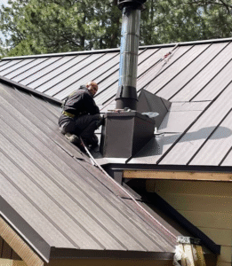 A worker wearing safety gear is installing or repairing a metal chimney pipe on a sloped metal roof.
