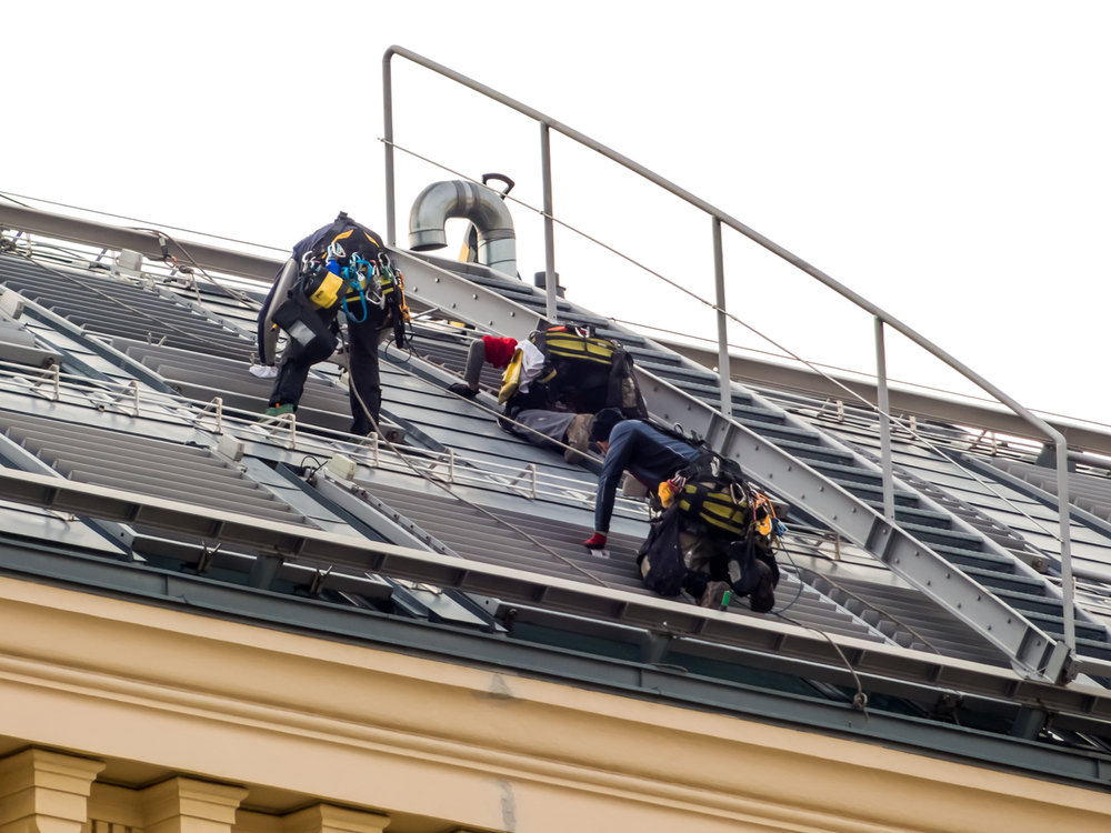 Three workers wearing safety gear are installing or maintaining equipment on a sloped metal roof with railings.