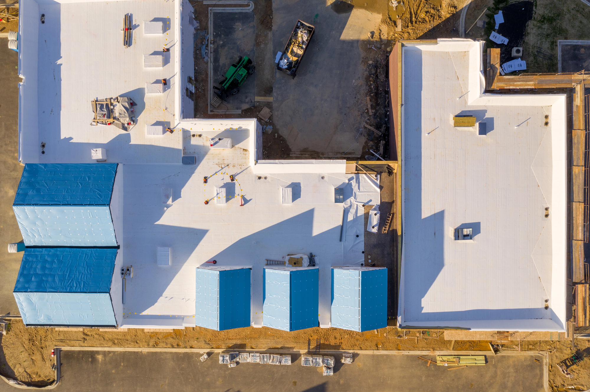 Aerial view of a construction site with white roofing, blue-roofed sections, construction equipment, and building materials visible around the structure.