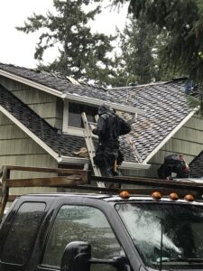 A worker in rain gear stands on a ladder, repairing the roof of a house on a rainy day, with tools and equipment visible nearby.