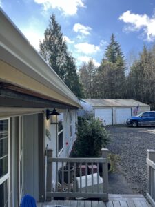 A view from a porch showing the side of a house, a gravel driveway, a blue truck, two garages, an American flag, and trees under a partly cloudy sky.
