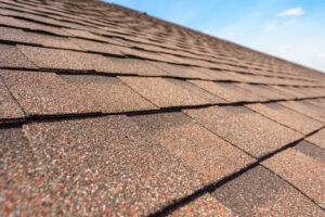 Close-up view of brown asphalt roof shingles on a sloped roof, with blue sky visible in the background.