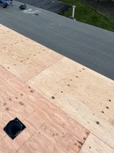 Plywood sheets installed on a roof, with a pipe protruding through one section; tools and a partially covered roof visible in the background.