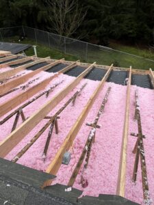 Partially constructed roof with exposed wooden beams and pink insulation material between them; trees and a fence are visible in the background.