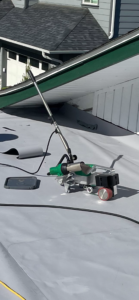 A handheld heat welding tool and other equipment rest on a flat, gray rooftop near a house with green and white trim on a sunny day.