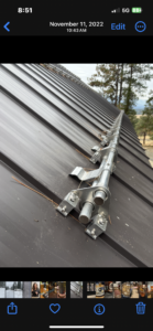 A close-up view of a metal snow guard bar installed along the ridge of a brown metal roof, with trees visible in the background.