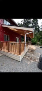 A covered wooden deck with a railing extends from a red house, surrounded by gravel and trees in the background.