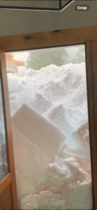 A doorway opens to a massive pile of snow blocking the entrance, with large snow slabs and outdoor structures partially visible beyond.